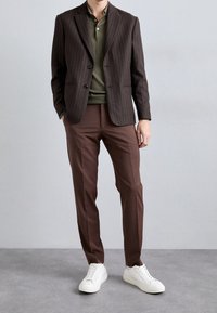 Brauner Anzugblazer mit feinen Streifen über einem grünen Poloshirt, kombiniert mit braunen Hosen und weißen Sneakern. Der Blazer hat zwei Knöpfe und einen Reverskragen.