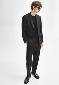 Zwarte op maat gemaakte suit met een blazer met één knoop en rechte broek. Draag het over een zwart T-shirt, gecombineerd met glanzende zwarte schoenen.