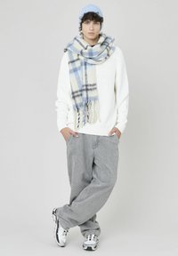 Pull en tricot blanc, pantalon gris large, et bonnet bleu. L'écharpe présente un motif tartan en bleu, crème et noir avec des franges.