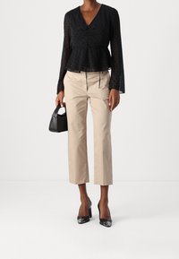 Donna che indossa una blusa nera trasparente a pois con maniche lunghe, pantaloni beige a vita alta, tacchi neri e tiene in mano una piccola borsa nera.