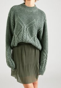 Ylisuuri vihreä neulottu paita, jossa on letiköityjä kuvioita, yhdistetty laskostettuun oliivinvihreään hameeseen. Pehmeä pinta ja rento istuvuus näkyvät.