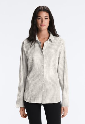 Femeie cu păr lung și închis la culoare, purtând o cămașă bej butonată cu un model fin în carouri și pantaloni negri, cu fața spre înainte pe un fundal simplu.