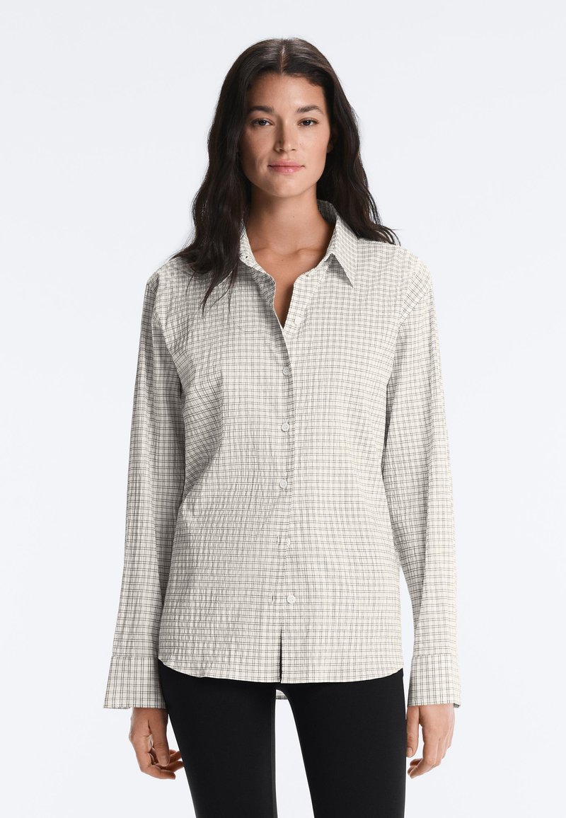 Femme aux longs cheveux foncés portant une chemise beige boutonnée avec un motif à fines carreaux et un pantalon noir, faisant face à un fond uni.
