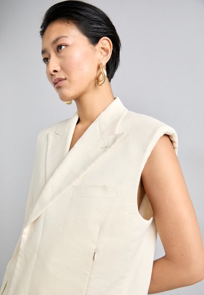 Blazer sans manches en lin beige avec un col cranté, une poche frontale et une coupe ajustée. Modèle portant de grandes boucles d'oreilles en or.