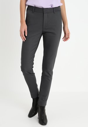 Nainen pukeutuneena kapealahkeisiin tummanharmaisiin housuihin ja mustiin nilkkureihin, seisomassa yksiväristä valkoista taustaa vasten.