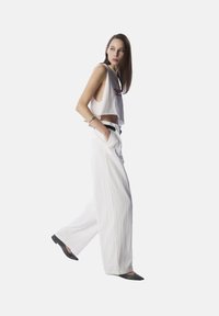 Top corto bianco con testo rosso, abbinato a pantaloni bianchi larghi. La modella indossa scarpe grigie e ha lunghi capelli castani lisci.