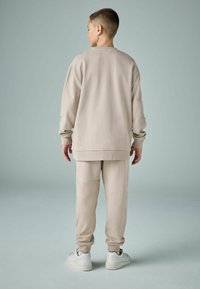 Bežs pulovers un atbilstošas bikses no mīksta auduma; brīva piegriezuma ar ribotiem piedurknēm un apakšmalu. Stilizēts ar baltām sporta kedām.