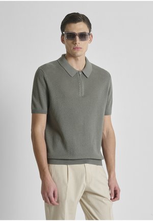 Hombre con polo gris de manga corta de punto con cuello con cremallera y pantalones beig plisados, con gafas de sol oscuras, de pie sobre un fondo neutro.