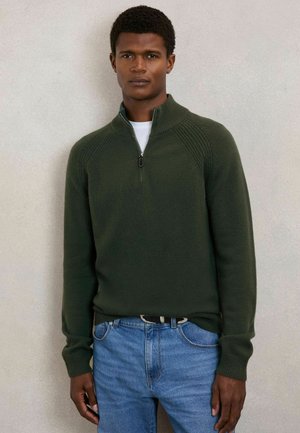 Sötétzöld cipzáras pulóver bordás díszítéssel és hosszú ujjakkal, kék farmernadrággal párosítva, egyszerű nyakkivágással és texturált részletekkel.