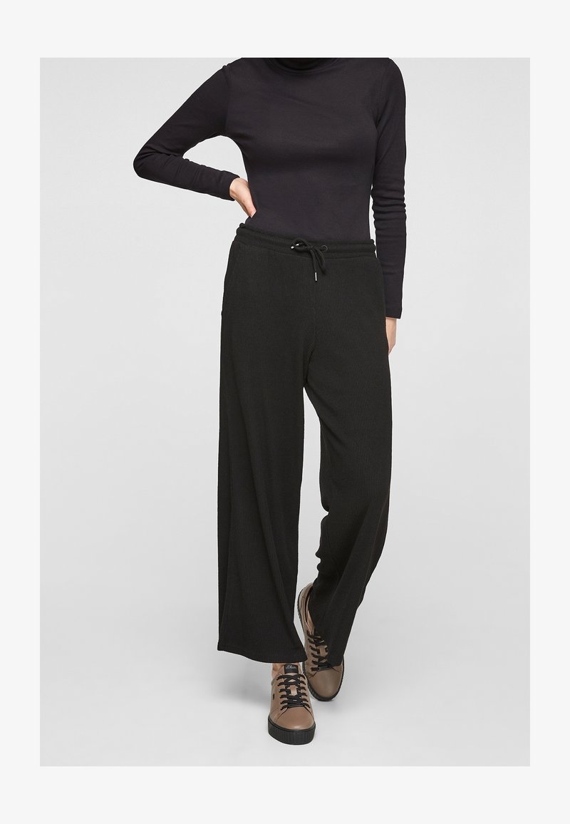 Personne portant un pantalon noir ample côtelé avec taille à cordon, un haut noir à manches longues et des baskets marron à lacets, debout sur un fond gris.