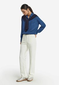 Hemd aus blauen Satin mit langen Ärmeln, kombiniert mit einem dunkelblauen Pullover um den Hals, dazu lockere off-white Hosen und metallisch silberne Sneakers.