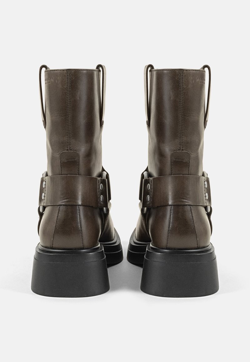 Braune Leder-Stiefeletten mit glatter Oberfläche, breiten schwarzen Gummisohlen und einem dekorativen Riemen mit metallicfarbigen Nieten am Knöchel.