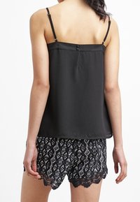 Svart satin camisole med justerbara band och knappdetalj på baksidan; bärs med svarta mönstrade shorts med spetskant.