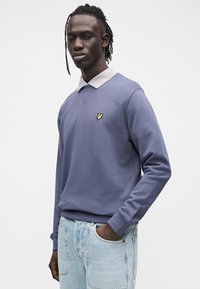 Lilla pusa heledatehallide kraega, mis on rinnal väikese kollase logoga. Kanditud koos helesiniste teksapükstega.