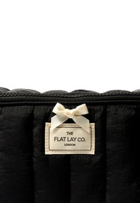 Funda acolchada negra con tejido texturizado, que presenta un cierre superior, una etiqueta color crema con el nombre de la marca y un pequeño lazo decorativo.