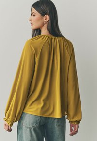 Långärmad blus i senapsgul, med en rynkad halsringning och elastiska ärmslut, tillverkad av mjukt, lätt material. Kombinerad med denim.