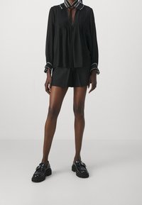 Blouse noire plissée avec une fermeture à nouer et un col à motifs, associée à un short noir et des mocassins noirs épais. Texture de tissu lisse.