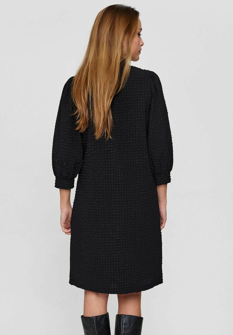 Czarna sukienka o fakturowym wykończeniu z rękawami 3/4, sięgająca do kolan, w luźnym kroju i z wzorzystą tkaniną. Brak widocznych elementów metalowych ani akcentów.