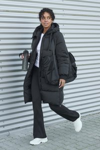 Melnā pūšanas jaka ar kapuci, komplektā ar baltu topu un melnām biksēm. Nēsā melnu ūdens pudeli un somu ar režģa tekstūru. Balti sporta apavi.