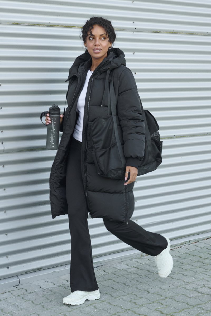 Melnā pūšanas jaka ar kapuci, komplektā ar baltu topu un melnām biksēm. Nēsā melnu ūdens pudeli un somu ar režģa tekstūru. Balti sporta apavi.