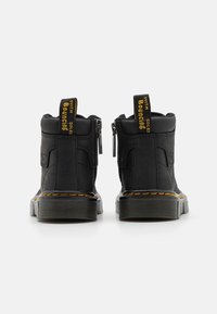 Černé kotníkové boty s texturovaným povrchem, bočními zipy a výraznými žlutými táhly. Mají silnou podrážku z gumy s výrazným vzorem.