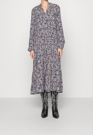 Frau trägt ein langärmliges, geblümtes Midikleid mit V-Ausschnitt und schwarze, bestickte Stiefel bis zur Mitte der Wade, steht vor einem schlichten Hintergrund.