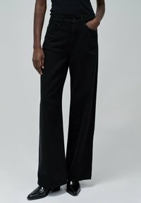 Jeans negros de pierna ancha con una cintura alta, con bolsillos y una textura de denim suave. Combinados con botas de tacón negro puntiagudas.