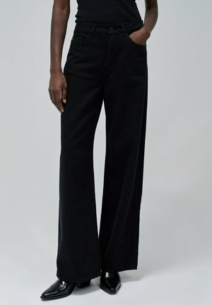Jeans negros de pierna ancha con una cintura alta, con bolsillos y una textura de denim suave. Combinados con botas de tacón negro puntiagudas.