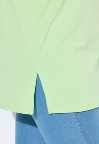 Hellgrünes Baumwoll-T-Shirt mit seitlichem Schlitzdetail; kombiniert mit hellblauen Jeans. Nahaufnahme des Saums und der Stoffstruktur.