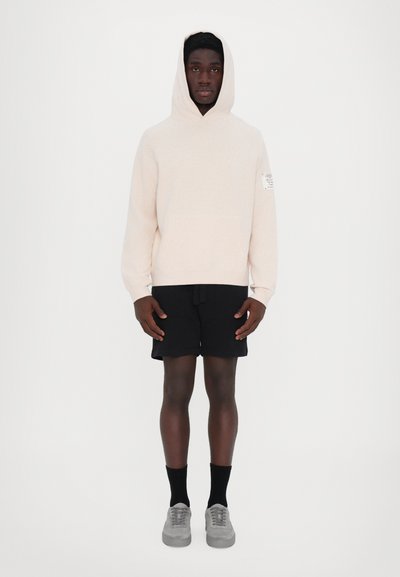 Light beige hoodie with a front pocket, paired with black shorts. Model wears gray sneakers and dark socks. Simple design with no patterns.