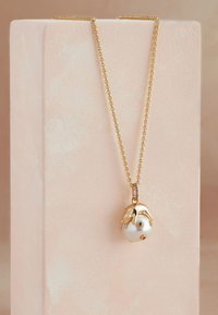 Zelta ķēdes kaklarota ar apaļu baltu kulonu, kas ietver zaļus un violetus akcentus. Kulonam ir teksturēts, organiskas formas dizains.