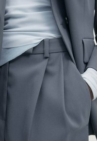 Grijze op maat gemaakte pak met een gestructureerde blazer en geplooide broek. Zachte blauwe blouse eronder, met een gladde stoftextuur en een getailleerd ontwerp.