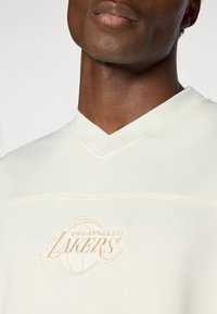 Kræmfaret sportsjersey i cremefarve med V-halsdesign, der har struktureret stof og et præget "Los Angeles Lakers" logo i matchende farve.