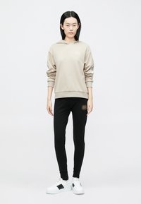 Μπεζ φούτερ με κουκούλα και λογότυπο στο στήθος, συνδυασμένο με μαύρο κολάν και λευκά αθλητικά παπούτσια με μαύρες λεπτομέρειες, σε απλό φόντο.