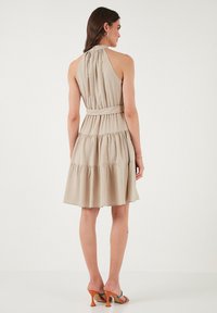Vaalea beige halterneck-mekko, jossa on istuva yläosa, rypytetty kaulus ja kerroksellinen hameosa. Mekossa on yhteensopiva vyö ja sileä pinta.