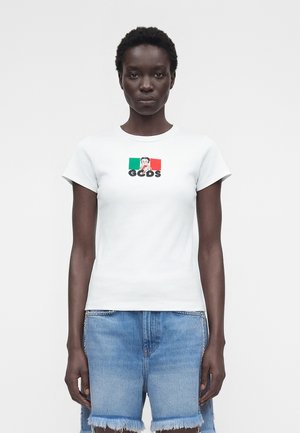 Kobieta w białym T-shircie z małą włoską flagą oraz postacią z kreskówki nad napisem „GCDS”, w zestawieniu z postrzępionymi, niebieskimi szortami jeansowymi.