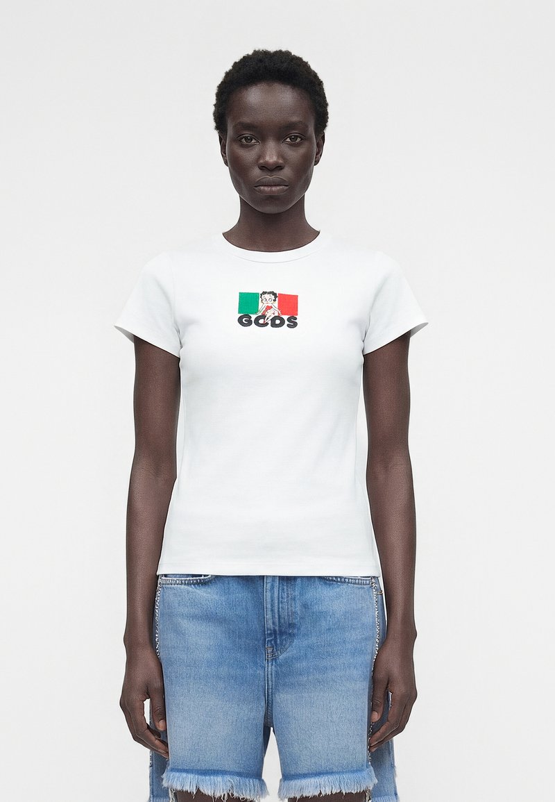 Γυναίκα που φοράει λευκό T-shirt με μικρή ιταλική σημαία και χαρακτήρα κινουμένων σχεδίων πάνω από το κείμενο «GCDS», συνδυασμένο με ξεφτισμένες μπλε τζιν σορτς.