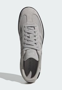 Zapatilla gris con materiales de ante y textil, cordones planos, puntera redonda y suela de goma negra con detalles de logo en la lengüeta.