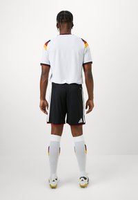 Biely futbalový dres s červenými a žltými akcentmi, čierne šortky s bielymi pruhmi, biele sieťované ponožky s čiernymi, červenými a žltými detailmi.