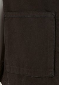 Ciemnobrązowy materiał o teksturowanym wykończeniu, z pojedynczą kieszenią naszywaną. Szwy proste z wykończeniem szytym. Materiał wydaje się trwały.