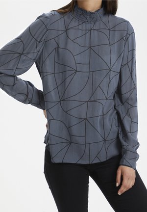 Femme portant un chemisier gris à manches longues avec des motifs abstraits en lignes noires et un col montant à volants, associé à un pantalon noir.