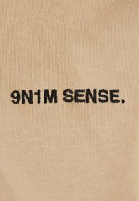 Il tessuto beige presenta un ricamo nero con la scritta "9N1M SENSE." Il materiale ha una texture liscia con un motivo a maglia fine.