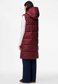 Mujer con un chaleco acolchado burdeos largo con capucha, camiseta de manga larga blanca, vaqueros de pierna ancha azul oscuro y zapatos marrones, de pie de espaldas.