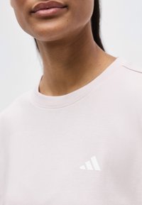 Gros plan sur une personne portant un T-shirt Adidas rose clair avec le logo visible sur la poitrine.