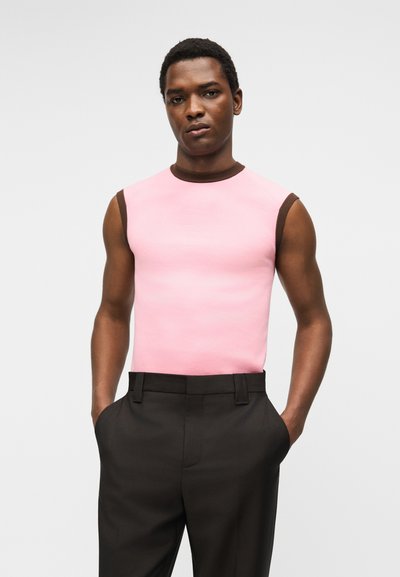 Άνδρας που φοράει ροζ αμάνικη μπλούζα με καφέ λεπτομέρειες και μαύρο παντελόνι, με τα χέρια στις τσέπες, μπροστά σε απλό φόντο.