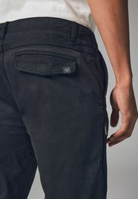 Fekete cargo nadrág hátulján logós zsebbekkel, oldalsó cipzáras zsebbel és textúrált anyaggal rendelkezik, modern dizájnt bemutatva.