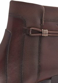 Botín de cuero marrón hasta el tobillo con panel texturizado y diseño elegante. Presenta un detalle decorativo de metal y costuras visibles.