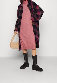 Veste chemise surdimensionnée à carreaux noirs et roses, robe en tricot rose, sac texturé beige et bottes à talons chunky noires.