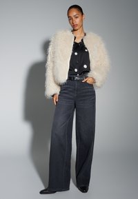 Nepbontjas in crème, zwart satijnen overhemd met witte stippen, wijde donkere jeans, zwarte riem en zwarte schoenen met puntige neus.