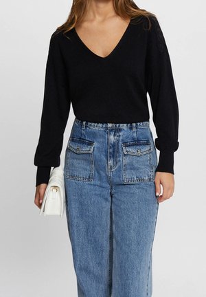 Mujer con un suéter negro de cuello en V y jeans de tiro alto azul con bolsillos delanteros, sosteniendo un bolso clutch blanco en una mano.